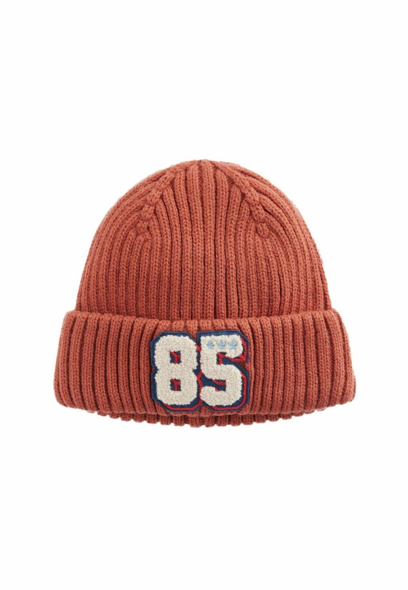 Stickad mössa i rostfärg, med ribbat mönster och uppvikt nederkant, prydd med en vit och blå broderad patch med texten '85'.