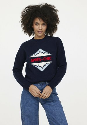 Marineblauer Strickpullover mit einem weißen und roten Berggrafik und dem Text "APRES-CHIC". Der Rundhalsausschnitt und die gerippten Bündchen ergänzen das Design.