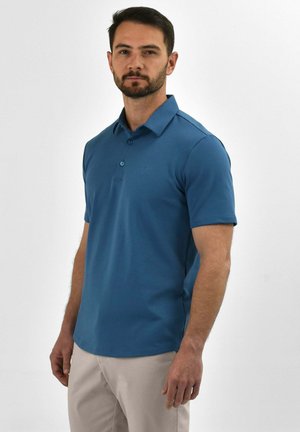 Man met baard, gekleed in een blauw poloshirt met korte mouwen en lichtbeige broek, staand tegen een effen witte achtergrond.