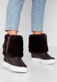 Botines impermeables marrones hasta el tobillo con un grueso puño de piel sintética, suela plataforma blanca con detalles texturizados, cierre con cremallera en el costado exterior.