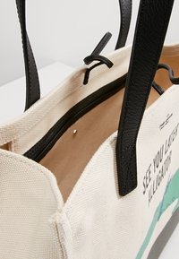 Borsa a tracolla in tela con manici in pelle nera, caratterizzata da un esterno color crema, una grafica stampata e una tasca interna con cerniera.