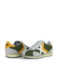 Zapatillas para hombre con un diseño en gamuza gris y verde oliva, acentos blancos, detalles amarillos y una suela de goma con una textura antideslizante.