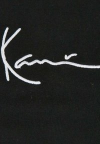 Svart stoff med fremtredende hvit brodert signaturdesign. Jevn tekstur med stingdetaljer, med fokus på flyten av bokstavene.