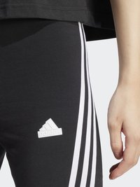 Leggings negre cu trei dungi albe pe laturi. Prezintă un logo Adidas alb pe coapsă. Material moale și elastic.