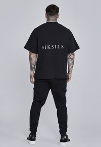 Crna majica kratkih rukava s natpisom "SIKSILK" u bijeloj boji na leđima, u kombinaciji s crnim cargo hlačama i crnim tenisicama.