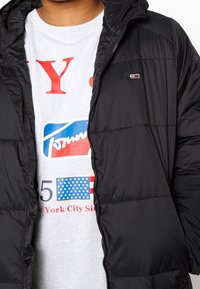 Giacca imbottita nera con cappuccio, dotata di chiusura a zip e una patch con logo. Sotto, una t-shirt grigia con grafiche rosse e blu.