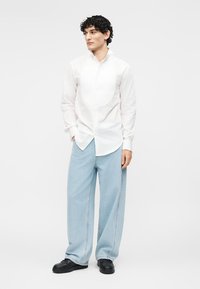 Άνδρας με σγουρά μαλλιά που φοράει λευκό πουκάμισο με κουμπιά, ανοιχτό μπλε παντελόνι με φαρδιές παντελόνες και μαύρα παπούτσια, στέκεται μπροστά σε λευκό φόντο.