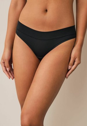 Nő fekete, varrás nélküli, közepesen magas bikinialsót visel, deréktól combközépig látható, semleges háttér előtt.