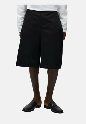 Persona che indossa pantaloncini neri a gamba larga fino al ginocchio, un top bianco a maniche lunghe e scarpe slip-on nere in pelle su uno sfondo uniforme.