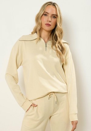 Vrouw met lang blond haar die een beige vest met rits en bijpassende broek met trekkoorden draagt, staand tegen een eenvoudige lichte achtergrond.