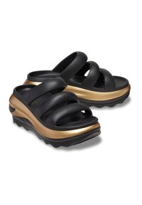 Zwarte sliders met drie gebogen bandjes, een gestructureerde zool en een gouden accent op een stevige zool met een golvende rand.