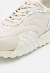 Gros plan d'une basket en cuir beige avec des lacets blancs et une semelle en caoutchouc blanc texturé montrant un motif de bande de roulement en forme de croix.