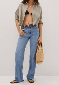 Camicia button-up marrone chiaro sopra un bikini nero, abbinata a jeans a gamba larga blu chiaro e una borsa a tracolla intrecciata con un bordo decorato.