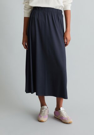 Femme portant une jupe midi bleu marine avec une taille élastique et des baskets roses et beiges, debout devant un fond clair uni.