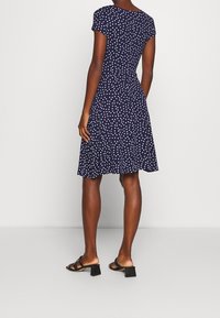 Kvinna klädd i en marinblå knälång klänning med vita prickar och svarta klacksandaler, stående mot en enfärgad bakgrund.