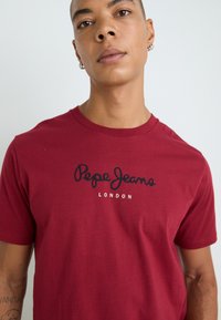 Κεραμιδί βαμβακερό T-shirt με κοντά μανίκια, διαθέτει εκτυπωμένο στο μπροστινό μέρος το λογότυπο "Pepe Jeans London" σε μαύρο χρώμα. Απαλή υφή, κλασική στρογγυλή λαιμόκοψη.