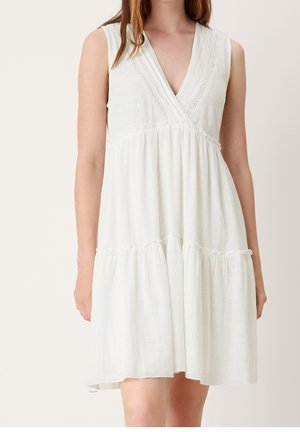 Woman wearing a sleeveless white dress with a V-neck and gathered skirt, standing against a plain light background.