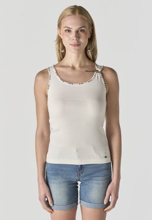 Mujer rubia con una blusa blanca sin mangas con detalle de tachuelas y shorts vaqueros azules remangados, de pie frente a un fondo liso.