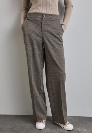 Woman wearing brown checkered wide-leg trousers, beige ribbed sweater, and beige canvas sneakers standing with hands in pockets.