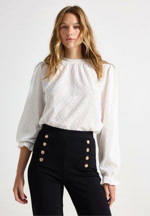 Blusa bianca ricamata con motivi a diamante, maniche a palloncino e dettagli in pizzo, abbinata a pantaloni neri a vita alta con bottoni dorati.