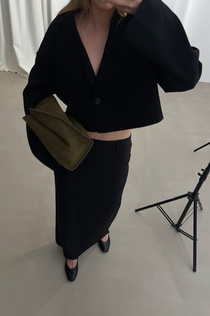 Black cropped jacket with long sleeves and a single button, paired with a black skirt and green suede clutch. Black pointed-toe shoes.
