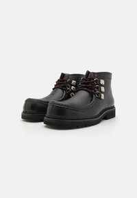 Bottes en cuir noir à bout rond, avec des détails de broderie décorative et des accents en métal argenté. Elles présentent des lacets rouges et noirs et une semelle robuste.