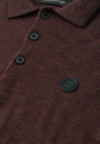 Polo en maille texturée marron avec des boutons noirs et un écusson rond noir près du bas à droite de la patte de boutonnage.