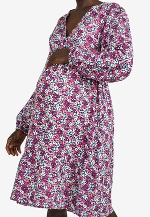 Personne enceinte portant une robe de maternité à manches longues avec un motif floral violet, rose et blanc, un décolleté en V et des poignets élastiques.