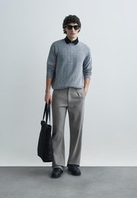 Camisola cinzenta com padrão, gola preta, calças cinzentas pregueadas, sapatos pretos e uma mala tote preta. O modelo tem cabelo encaracolado e usa óculos de sol.