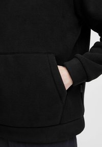 Svart fleecegenser med kengurulomme. Ribbestrikkede cuff og hem med en glatt tekstur. En liten merkeetikett er synlig.