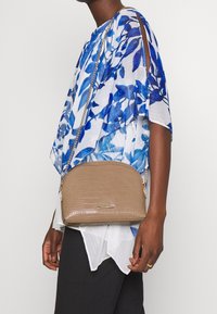 Hnědá crossbody taška s embosovaným krokodýlím vzorem, zaobleného tvaru, se zlatým řetízkem a minimalistickým kovovým detailem na přední straně.