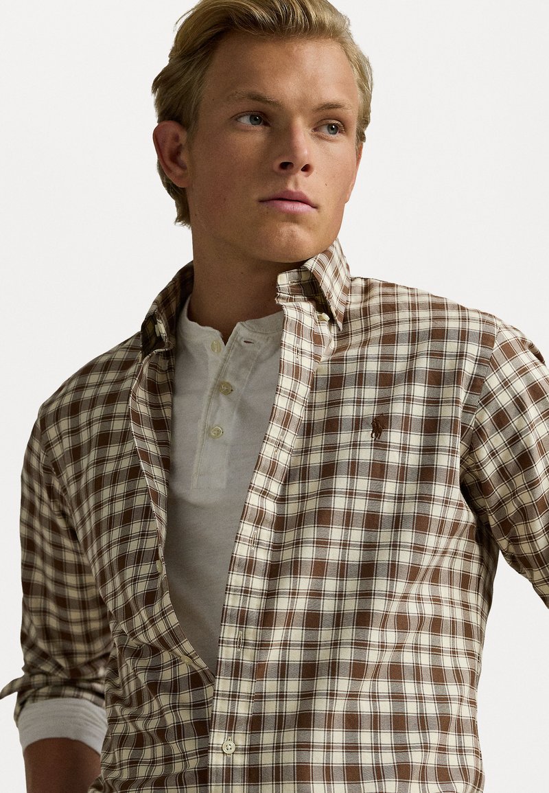 Camisa a cuadros marrón y crema con cuello abotonado, superpuesta a un henley gris claro. Presenta un pequeño logo en el pecho.