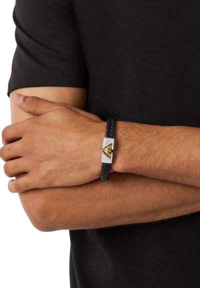 Homme portant un bracelet en cuir tressé noir avec un fermoir géométrique en argent et or, bras croisés sur une chemise noire à manches courtes.