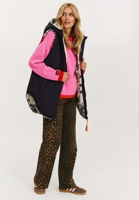 Eine Frau trägt einen pinken Pullover, eine schwarze ärmellose gesteppte Weste mit Leopardenmuster-Futter, Leopardenmuster-Hosen und braune Sneaker. Sie steht und schaut zur Seite.