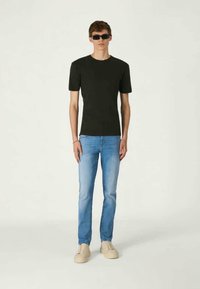Schwarzes, figurbetontes T-Shirt, hellblaue Slim-Jeans und beige Sneakers. Die Accessoires umfassen schwarze Sonnenbrillen. Das Model steht vor einem neutralen Hintergrund.