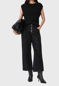Femme portant un haut noir sans manches texturé, un pantalon large noir taille haute avec des boutons, des bottes noires à talons et tenant un sac en cuir noir.