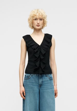 Femme aux cheveux blonds bouclés portant un haut noir sans manches avec un décolleté en V à volants et un jean large bleu, debout devant un fond blanc.