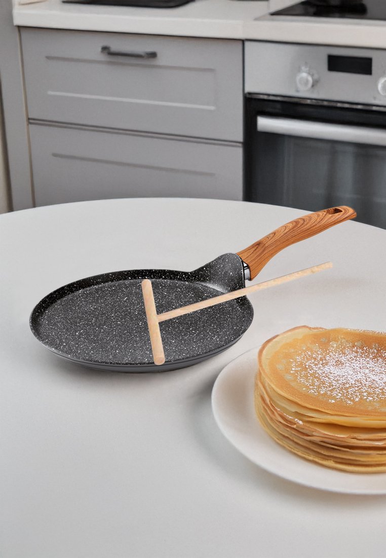 Poêle ronde noire avec un manche en bois, posée sur un plan de travail blanc. Une spatule en bois traverse la poêle ; une assiette de crêpes est à proximité.