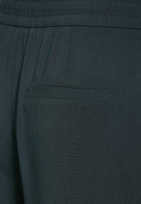 Zbliżenie na ciemne spodnie z tkaniny z elastycznym paskiem w talii i poziomą tylną kieszenią bez widocznego zapięcia.