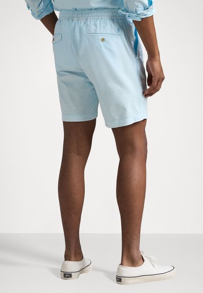 Un short bleu clair en tissu doux, avec une taille élastique et deux poches arrière avec fermetures à bouton. Des chaussures blanches décontractées complètent l'ensemble.