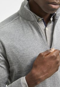 Grå zip-halströja i mjukt stickat material, med en rutmönstrad skjorta under, med knappdetaljer och en strukturerad kragn design.