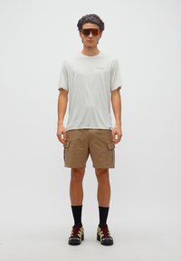 Νέος άνδρας στέκεται πρόσωπο προς τα εμπρός φορώντας μπεζ cargo βερμούδα, ανοιχτό γκρι μπλουζάκι Patagonia, μαύρες κάλτσες, παπούτσια για πεζοπορία και αθλητικά γυαλιά με κόκκινο φακό.