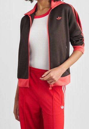 Person iført en sort og koralfarvet Adidas træningsjakke over en hvid top, kombineret med røde Adidas træningsbukser med hvide striber og logo.