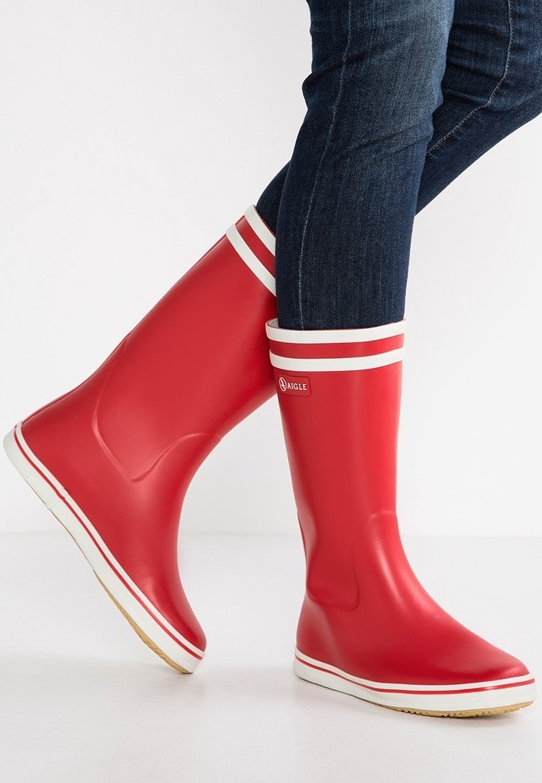 Personne portant des bottes en caoutchouc rouges avec des bandes blanches et un logo, associées à un jean denim bleu foncé, debout sur une surface blanche.