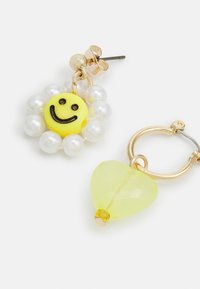 Charme de rosto sorridente amarelo com detalhes pretos, rodeado por pérolas brancas; combinado com um brinco de argola dourada com um charme de coração amarelo.