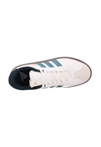 Zapatilla deportiva blanca con rayas color teal, que cuenta con una puntera de ante, forro textil y suela de goma marrón. Diseño clásico con un branding mínimo.