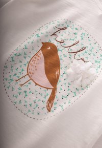 Bijela majica s ilustracijom smeđe ptice, natpisom "hello", zelenim akcentima, srebrnim zakovicama i detaljem bijelog cvijeta.