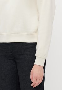 Kremfarget hettegenser med ribbestrikket halslinning og mansjetter, vist sammen med mørke denimjeans. Myk tekstur, avslappet passform.