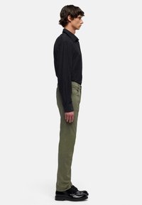 Camicia nera a maniche lunghe con chiusura a bottoni, pantaloni verde oliva e scarpe nere con lacci, vista di lato. Tessuto liscio, vestibilità su misura.