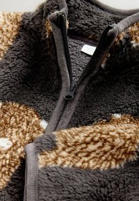 Fuzzy zip-up jacket in gray and brown, made from plush material. Features a black zipper and varying textures with spotted patterns.
